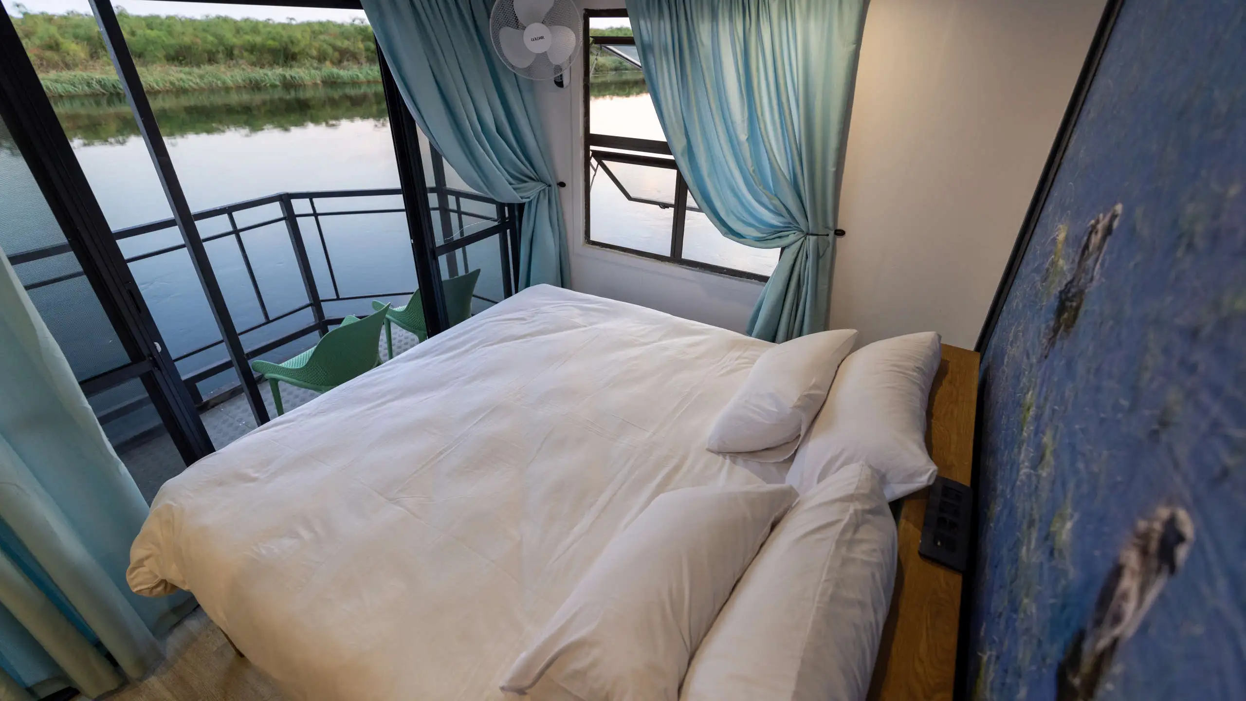 Cozy bedroom inside the Ngwezi houseboat in the Okavango Delta, with a view of the calm water through large windows, featuring soft blue curtains and a modern design.