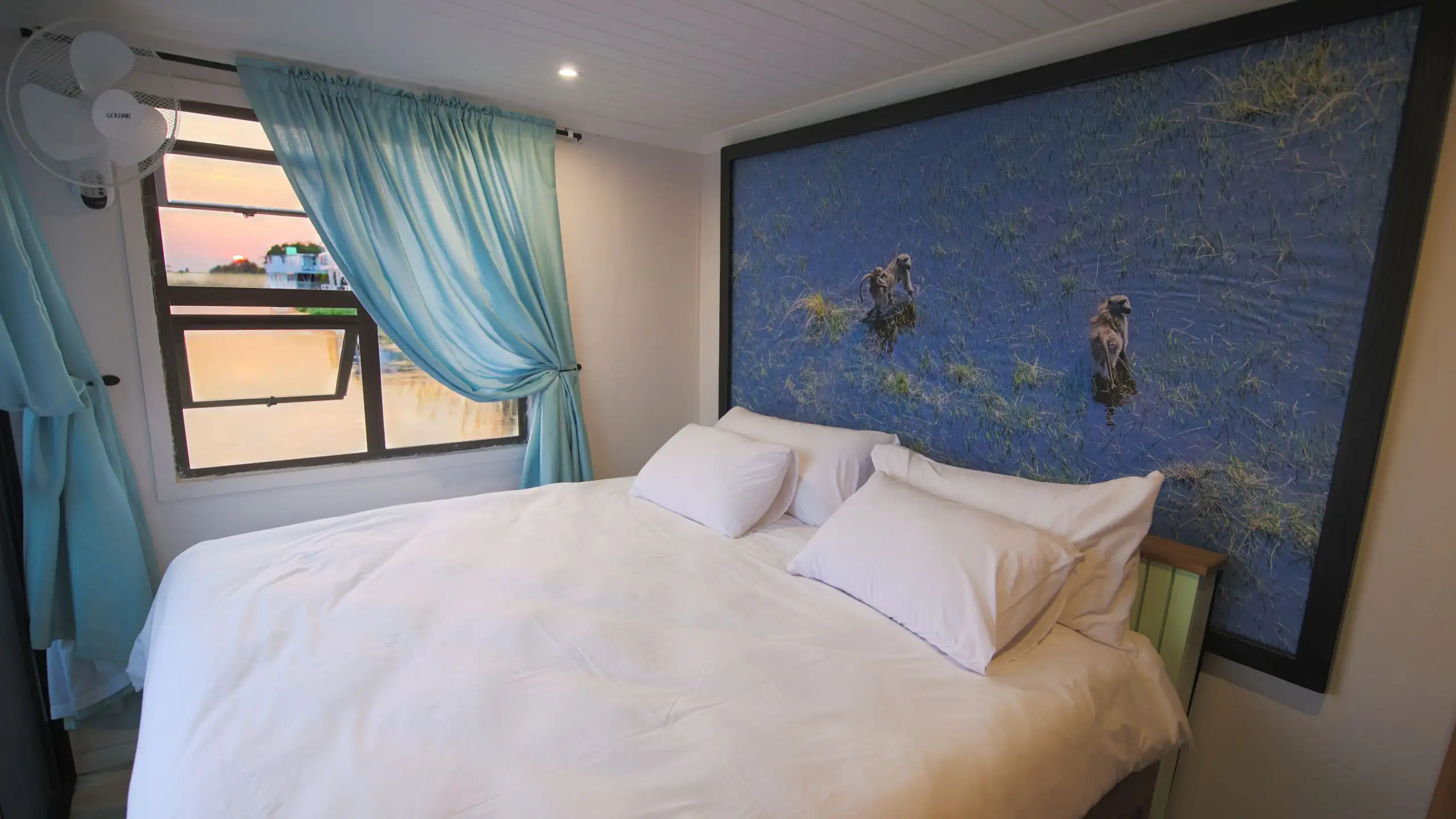 Ngwezi houseboat cabin featuring a comfortable bed with white bedding, framed by soft blue curtains and a beautiful wall art depicting monkeys in the Okavango Delta.