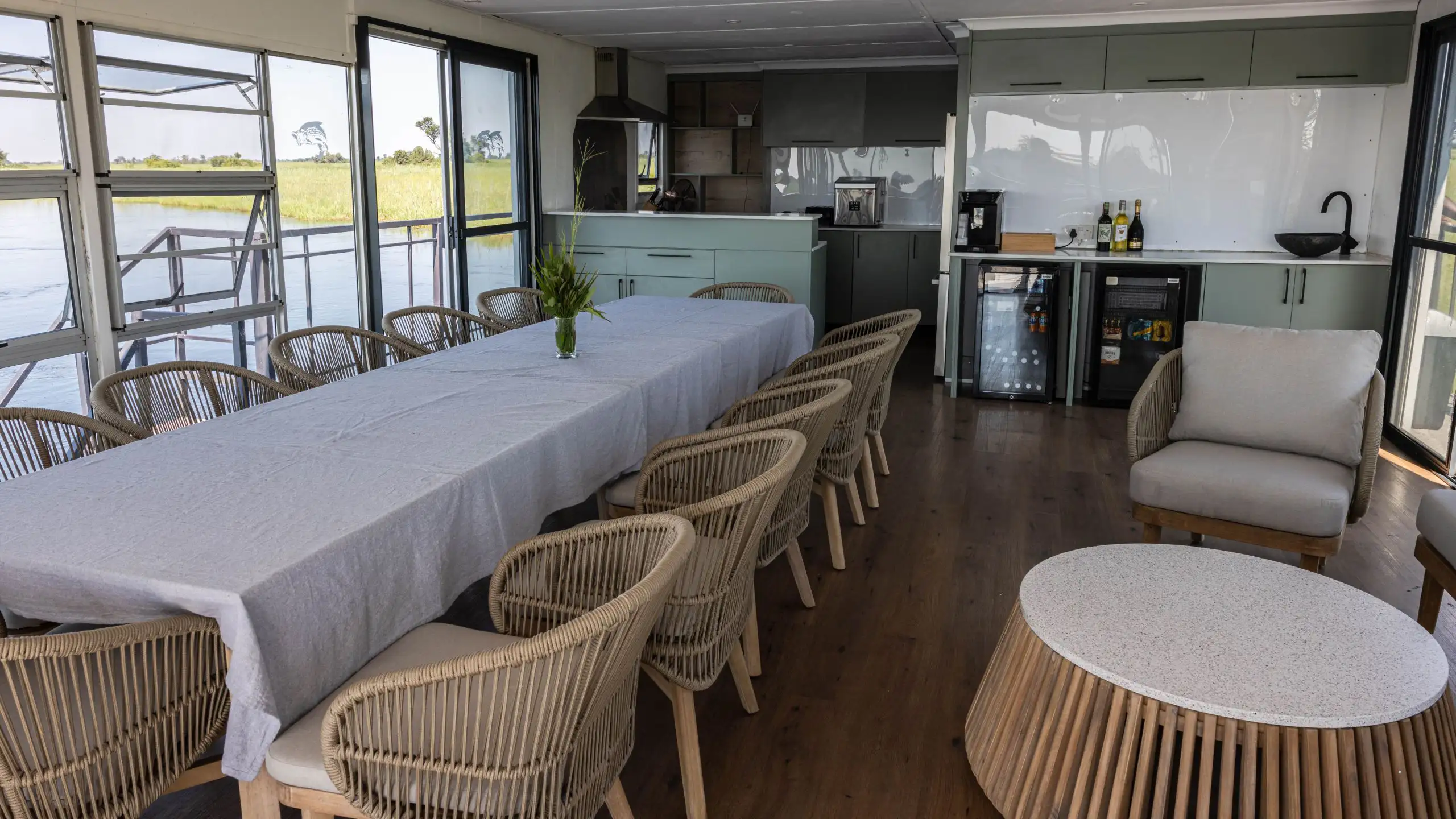 Interior view of the Ngwezi houseboat dining area, featuring a long table with wicker chairs, a modern kitchen, and large windows offering views of the Okavango Delta.