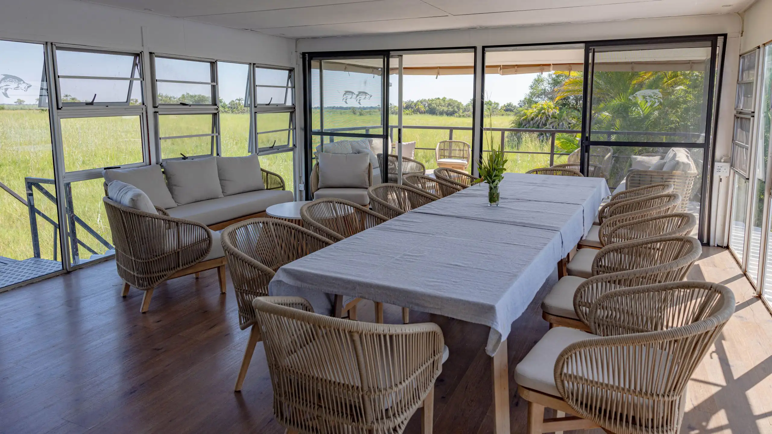 Interior view of the Ngwezi houseboat dining room featuring a long table surrounded by wicker chairs, with large windows offering scenic views of the Okavango Delta’s green landscape.â€