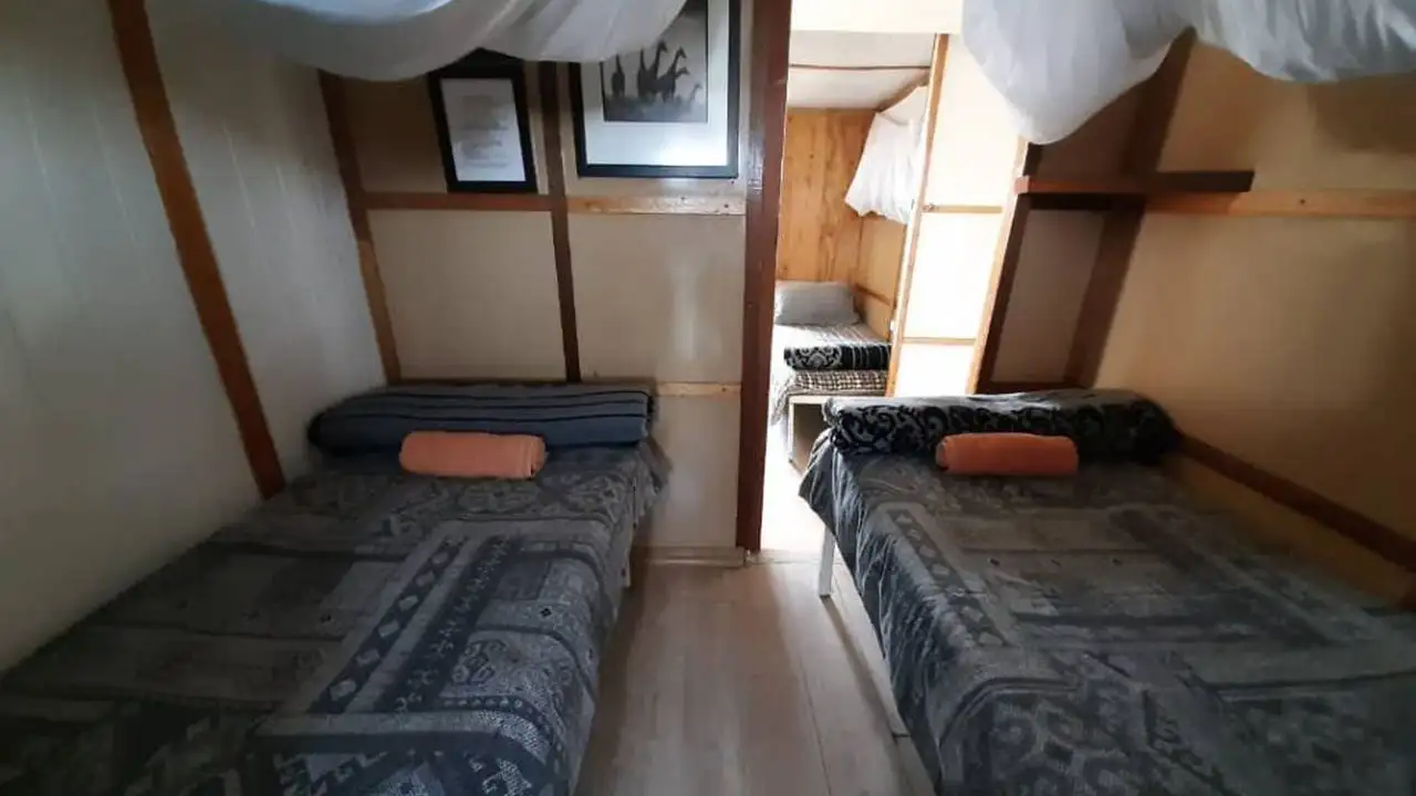 Interior of a twin bedroom on the Inkwazi Houseboat, showcasing two single beds with patterned bedding, rolled towels on the pillows, and framed artwork on the walls. A glimpse of a second room is visible through the door.