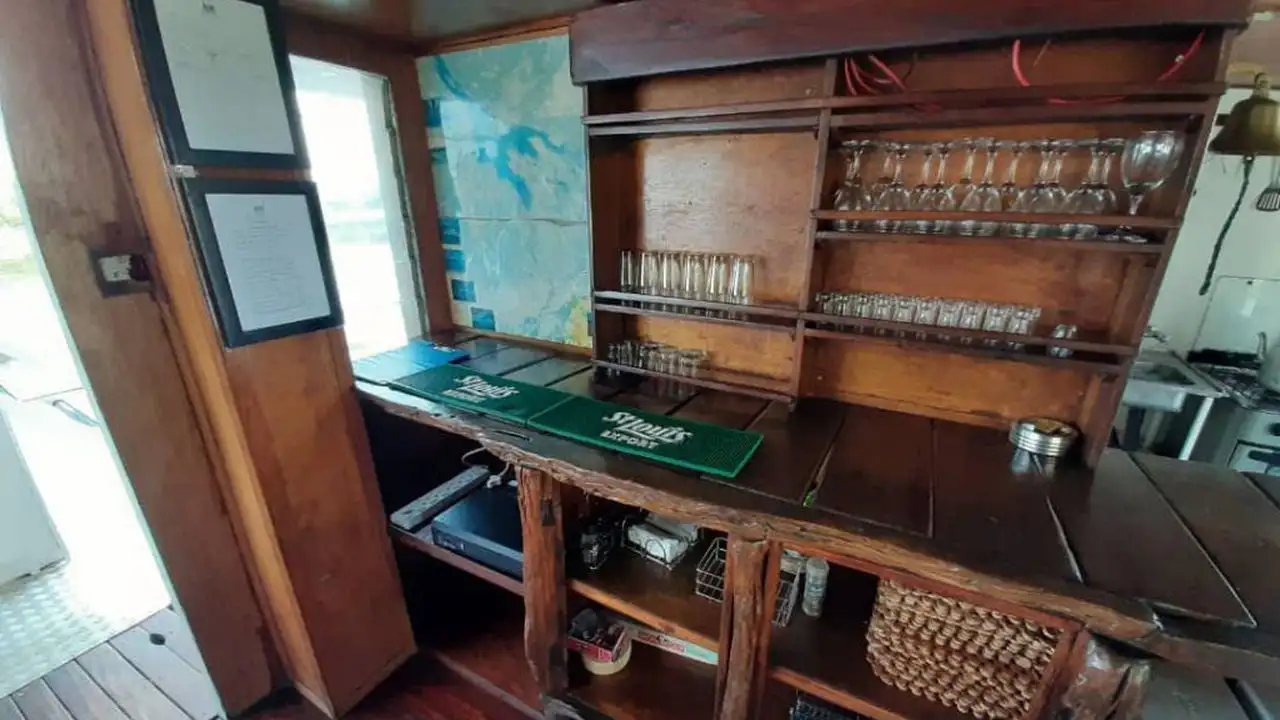 Interior view of the Inkwazi Houseboat's bar area, featuring a wooden bar counter with neatly arranged glasses and maps on the wall, creating a welcoming space for guests to relax and enjoy drinks on the Okavango Delta.