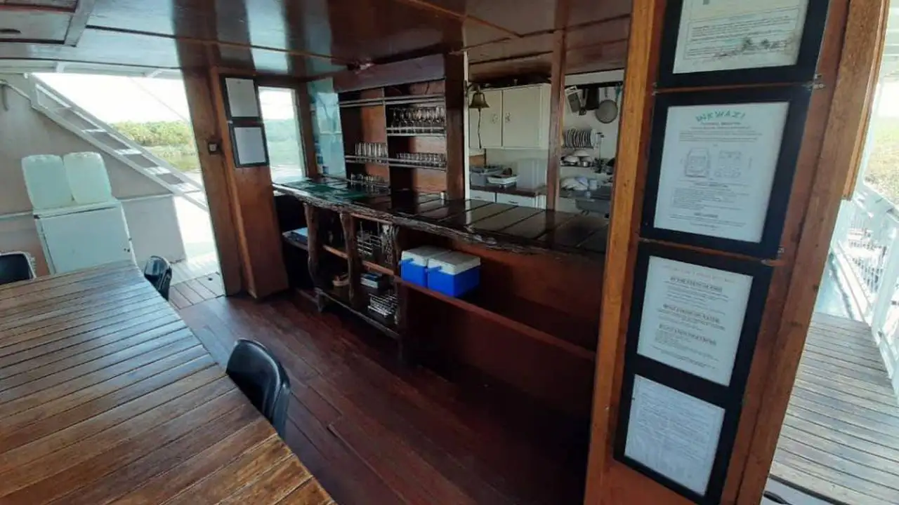 Interior view of the Inkwazi Houseboat bar area, showcasing a well-organized wooden bar counter with shelves stocked with glasses, a cooler, and framed documents on the wall, with a view of the kitchen area in the background.