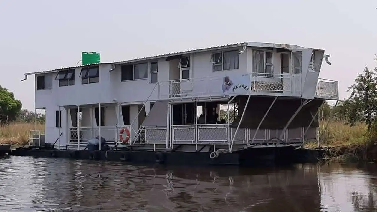 View of the Inkwazi Houseboat on the Okavango Delta, featuring the boat's exterior with a green water tank on the roof, a white structure with a large balcony, and a life ring visible on the side of the boat.