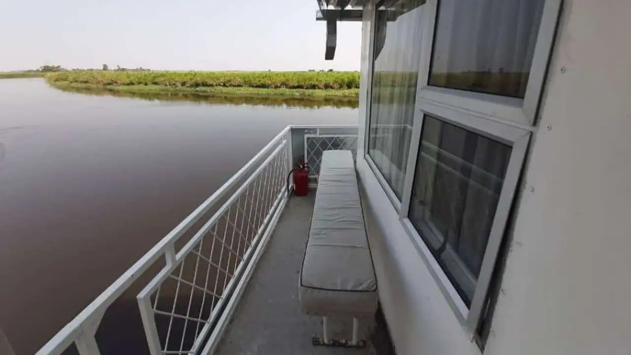 Exterior view of the Inkwazi Houseboat, showcasing a narrow walkway along the river with a bench and a fire extinguisher, offering a peaceful view of the surrounding riverbank and vegetation.