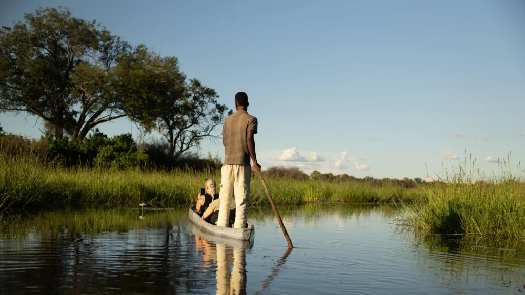 A Mokoro canoe with a guide poling through the calm waters of the Khwai River, surrounded by lush greenery and clear blue skies.