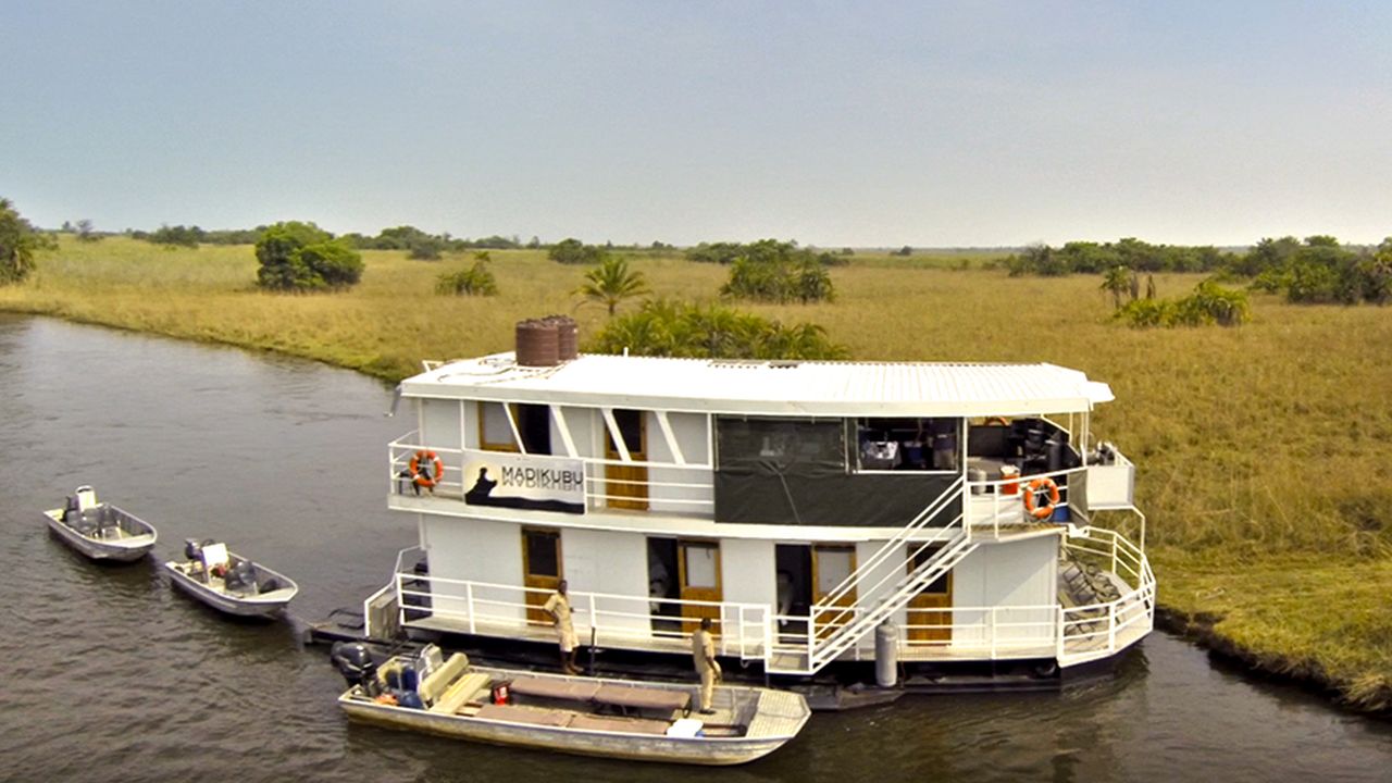 Aerial view of the Madikubu Houseboat cruising through the Okavango Delta, with small boats docked alongside, offering an immersive water-based safari experience.