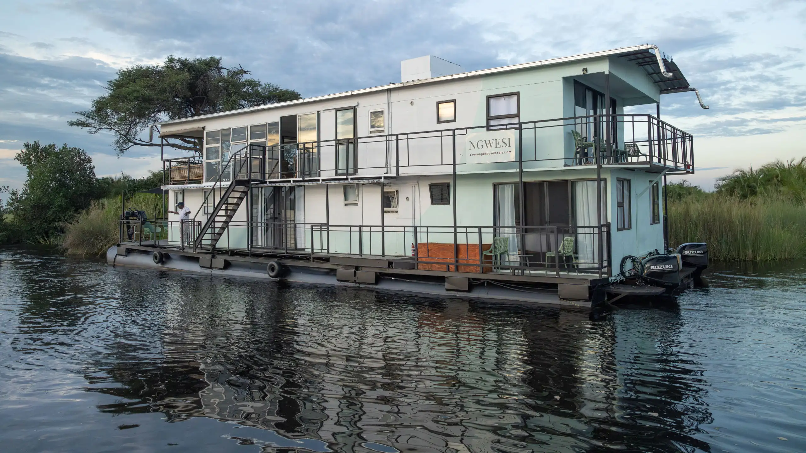 Ngwesi Houseboat floating on the calm waters of the Okavango Delta, showcasing its spacious two-story design with a staircase and modern furnishings, surrounded by lush vegetation and tranquil waters.