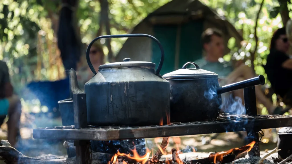 A kettle and cooking pots on a fire pit, steaming as they boil, with people sitting in the background at a camping site in the Okavango Delta.