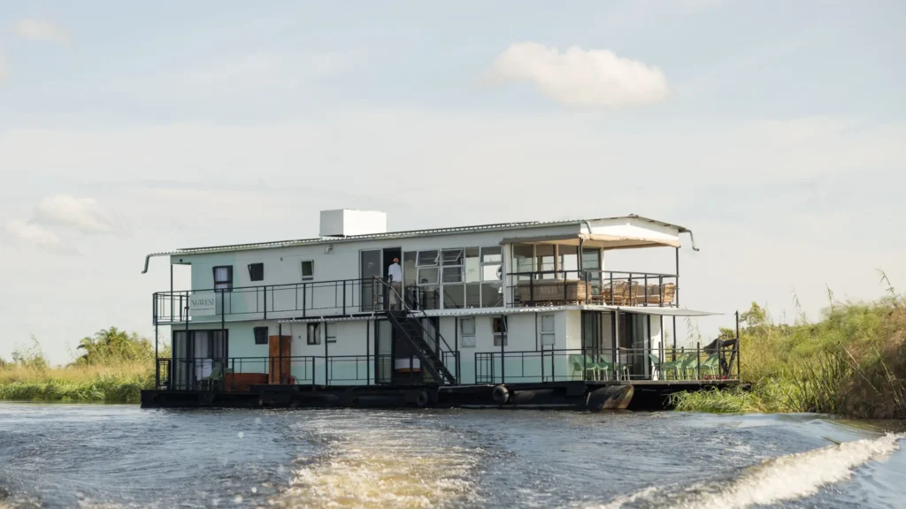 Ngwesi Houseboat cruising through the Okavango Delta, with clear skies, lush greenery along the waterâ€™s edge, and a person visible on the deck.