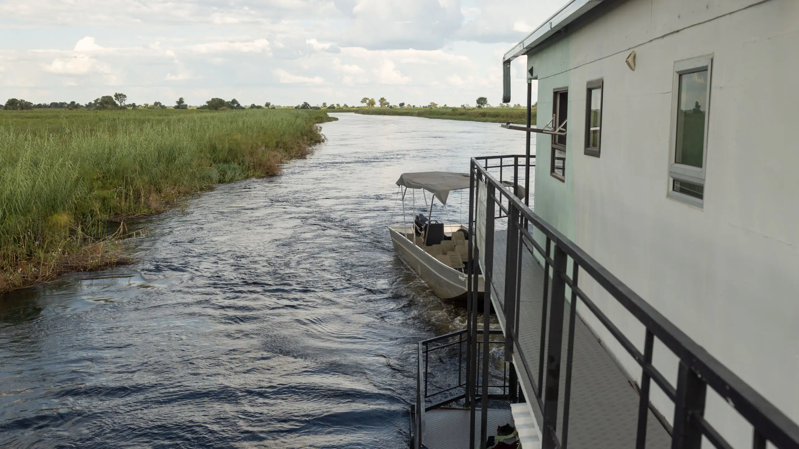 Scenic view of the Okavango Delta from a houseboat, with a small boat trailing behind, navigating through the winding waterways and lush green banks.